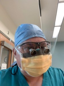 A close-up portrait of Dr. Rosenthal in a clinical setting. The individual is wearing teal scrubs, a light blue surgical cap, and a yellow medical face mask.
